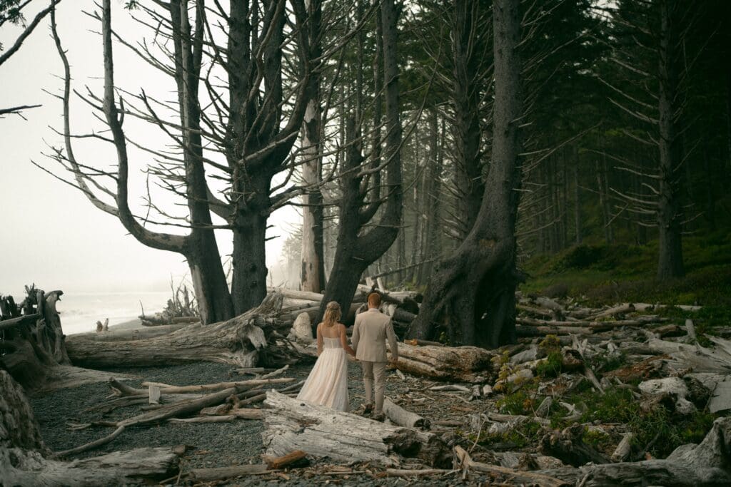 Bride and groom walk hand in hand through a driftwood‑covered Pacific Northwest beach forest on a foggy elopement day.