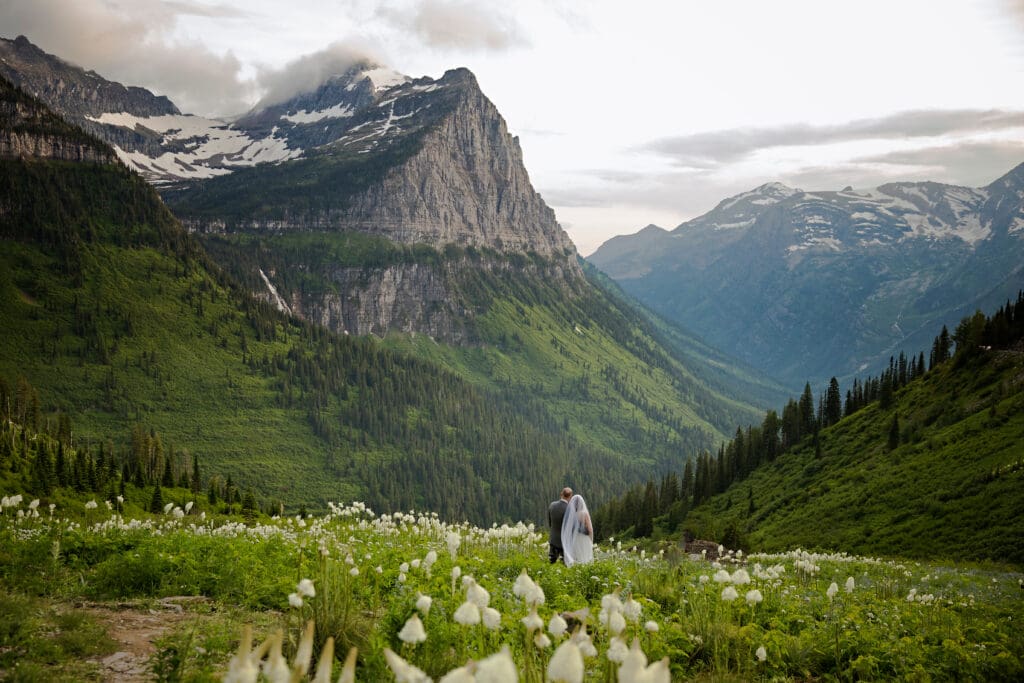 Eloping couple stands in a wildflower‑filled alpine meadow surrounded by dramatic Pacific Northwest mountains and evergreen valleys.