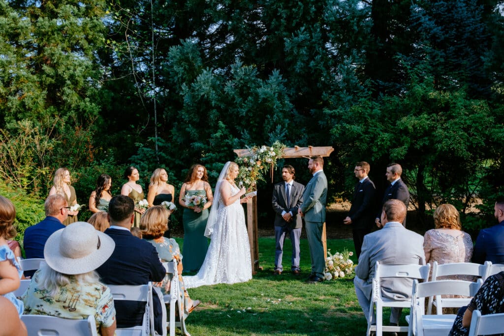 Outdoor wedding ceremony with a couple standing under a wooden arch, wedding party on each side, and guests seated on white chairs on a lawn