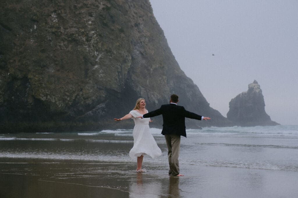 Couple eloping at Cannon Beach in Oregon holding out their arms by the ocean at low tide