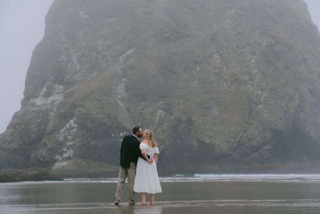 Bride and groom standing together in front of massive sea stack at Cannon Beach Oregon elopement