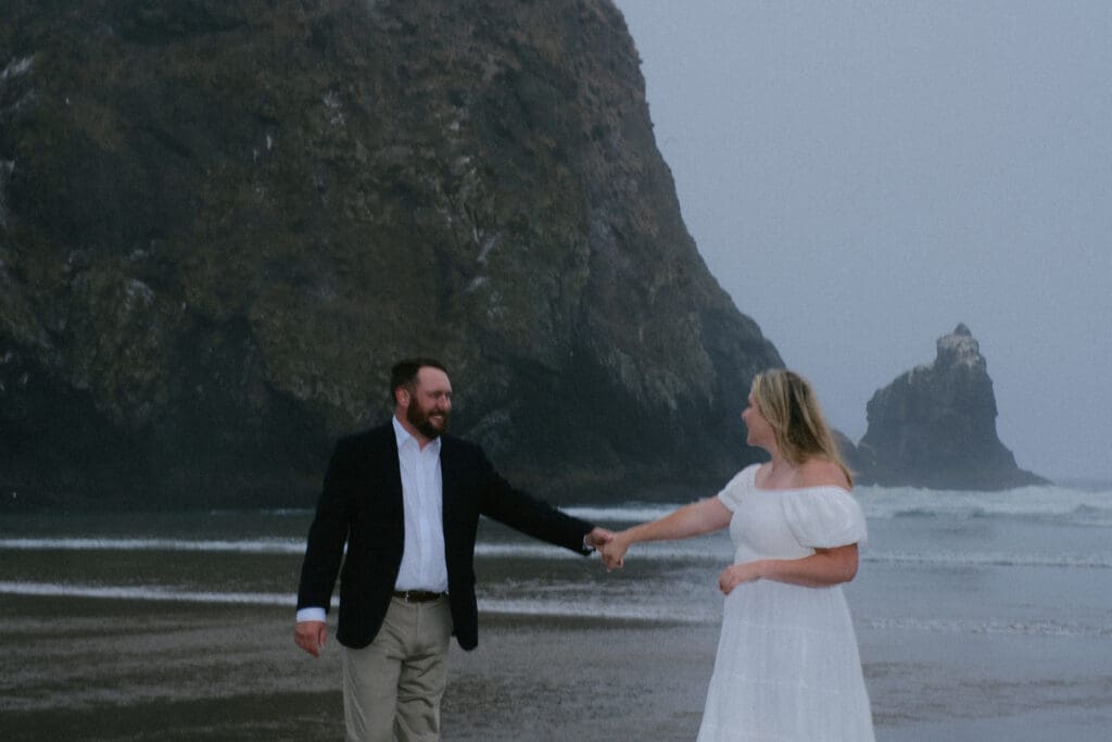 Bride and groom holding hands and kissing in front of rocky cliffs during a Cannon Beach elopement.