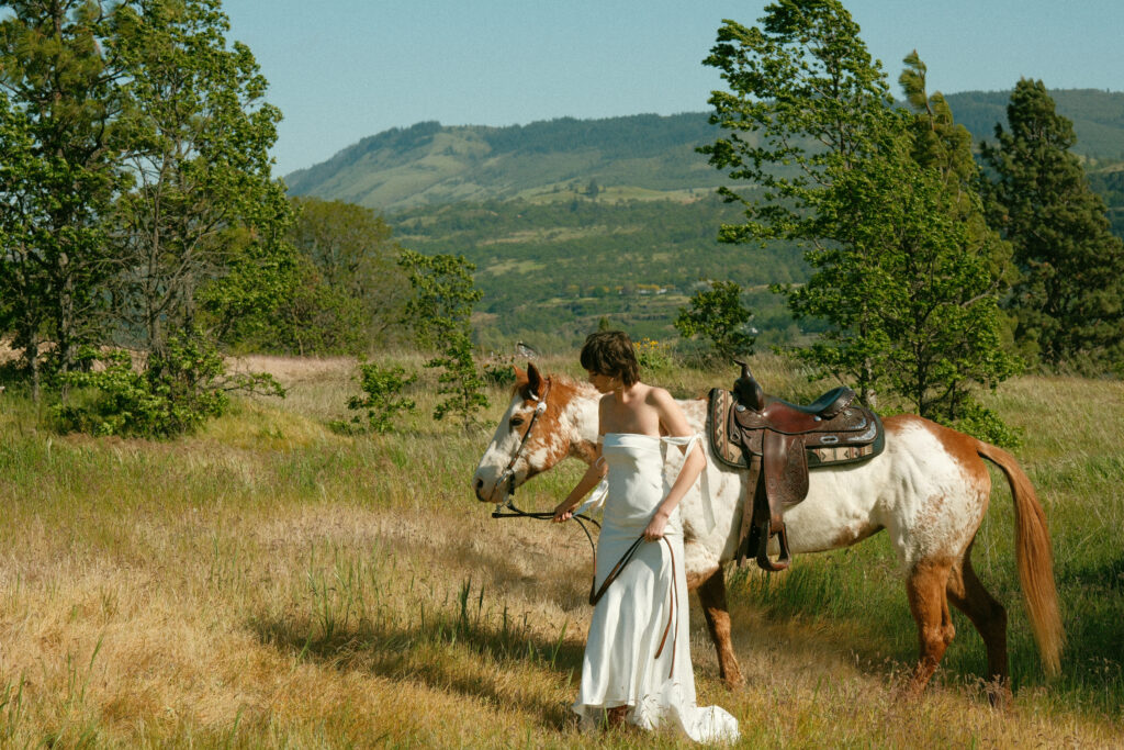Bride in simple white gown walking her horse during a Columbia River Gorge elopement in a sunlit meadow


