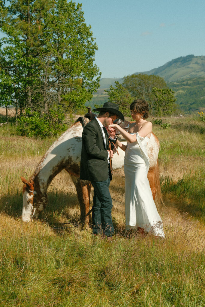 Columbia River Gorge elopement groom kissing bride's hand with their horse in a golden field with hills in the background
