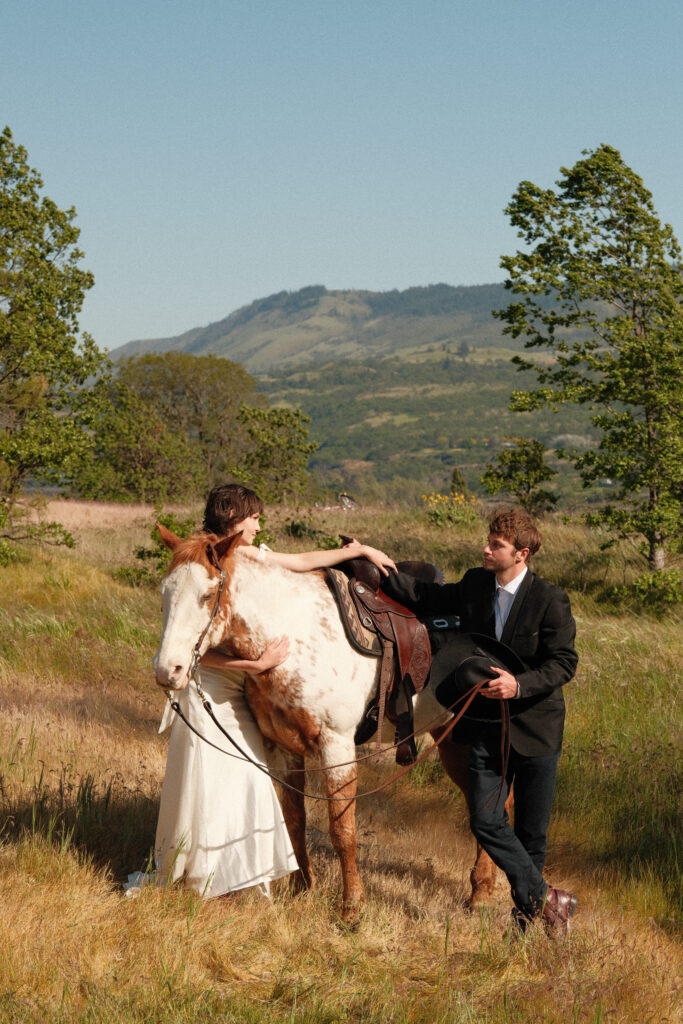 Bride hugging her horse while groom rests his hand on the saddle with Columbia River Gorge hills in the background

