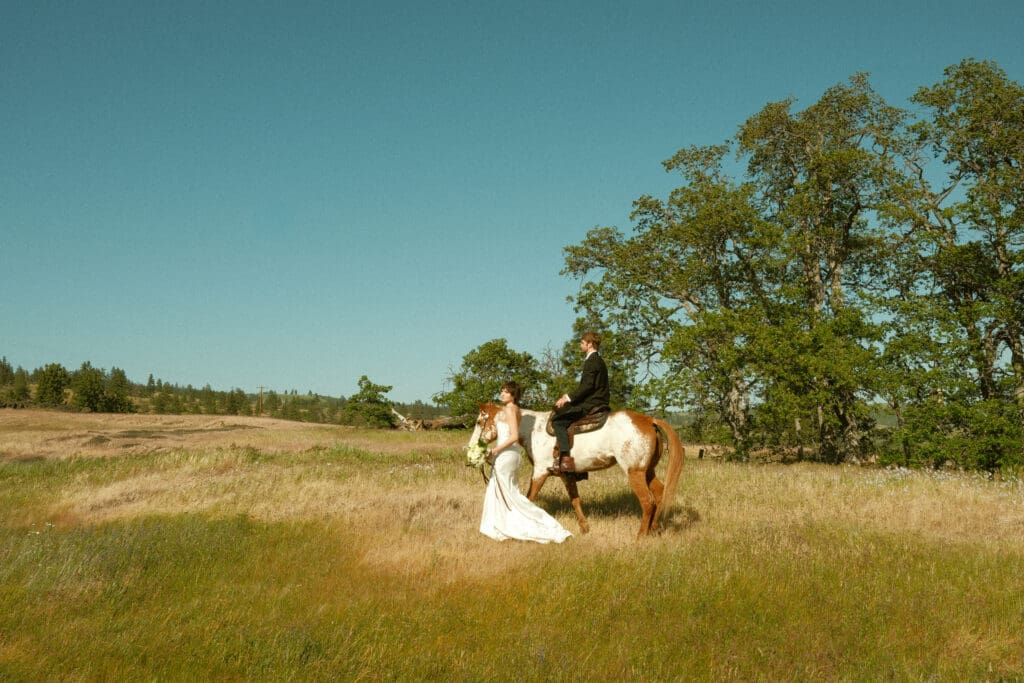 Bride leading a horse with her partner riding behind her in a sunlit Columbia River Gorge meadow

