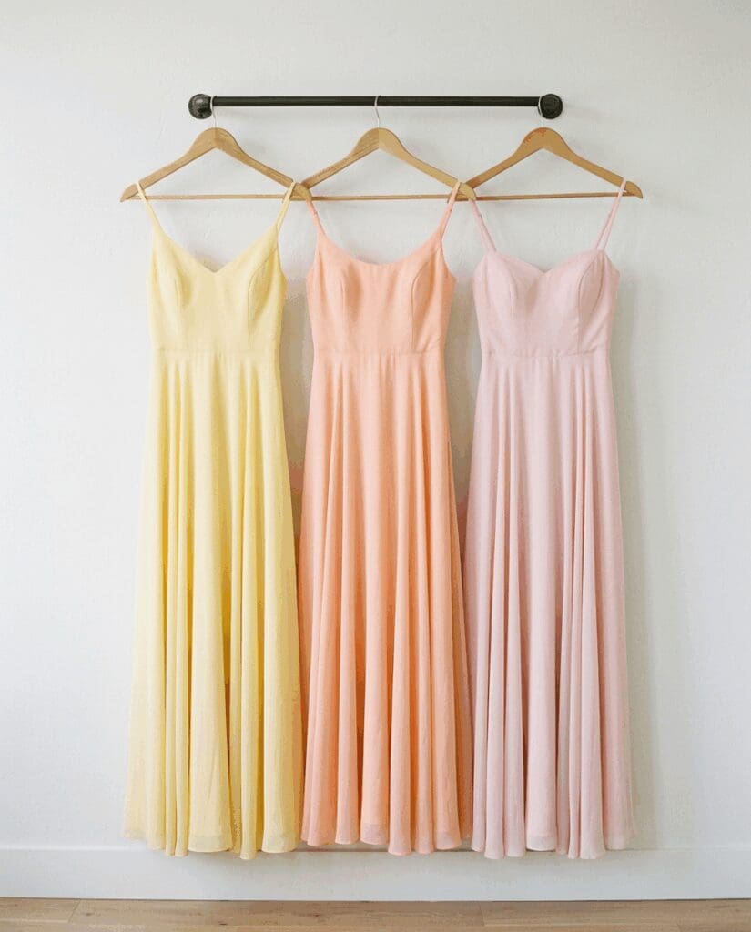 Trio of flowing bridesmaid dresses in PNW inspired 2026 sorbet wedding colors in pastel yellow, peach, and blush, hanging on a minimalist wall rod to showcase a soft sunset palette.

