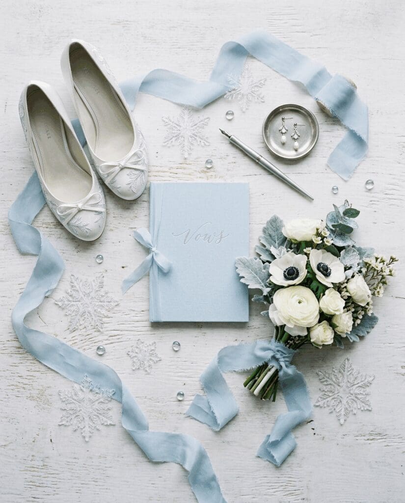 Winter bridal flatlay for a PNW wedding showing ivory flats, a dusty blue 2026 wedding vow book, silk ribbon, silver ring dish, and bouquet of white anemones and roses with dusty miller on a snowflake-textured backdrop.

