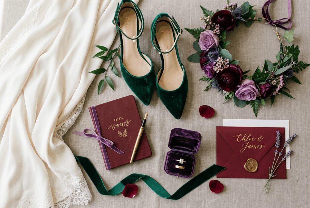 Flatlay of PNW fall wedding details with emerald heels, plum floral crown, vow book, invitation suite, and ring box styled in rich jewel tone 2026 wedding colors on a soft fabric backdrop.

