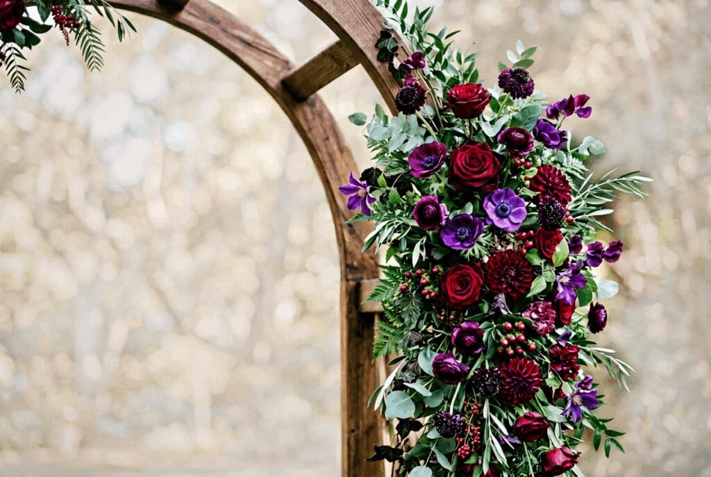 Romantic fall ceremony arch at a PNW wedding covered in jewel toned 2026 florals including deep red roses, burgundy blooms, purple ranunculus, and lush greenery climbing a wooden frame.

