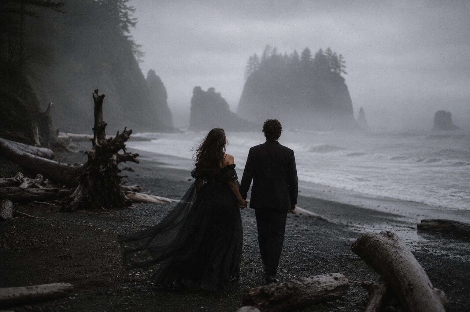 A couple in dark romantic attire walks along the misty shoreline at Rialto Beach in a Twilight-inspired Olympic National Park wedding scene with dramatic driftwood, dark water, fog, and overcast skies.