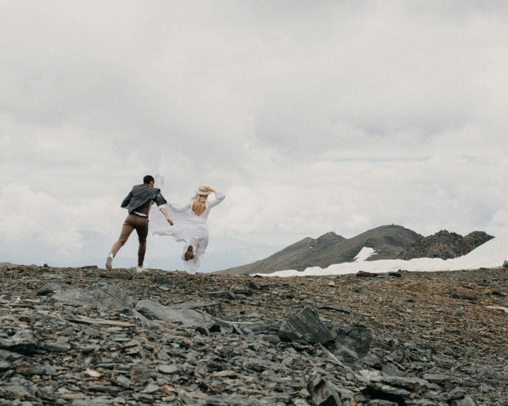 Couple running across a mountain landscape during changing weather conditions, highlighting the importance of planning for PNW elopement weather.