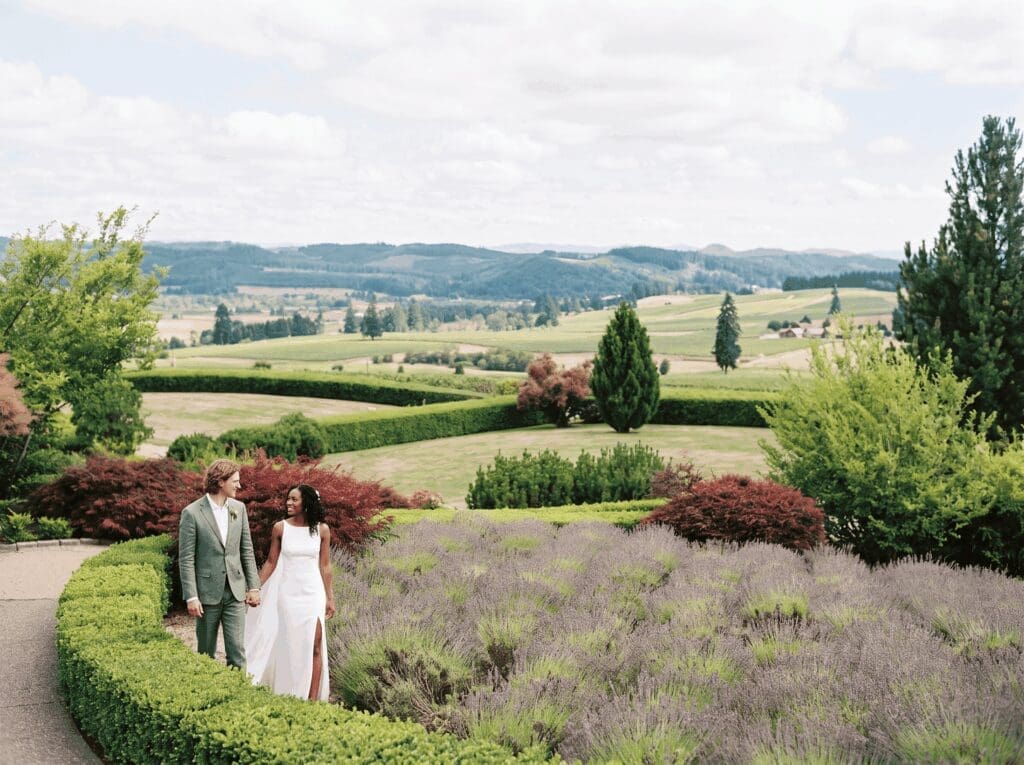 King Estate wedding couple holding hands along the lavender garden path, with panoramic vineyard and farmland views just outside Eugene in the Southern Willamette Valley.