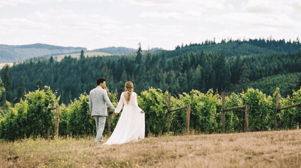 Bride and groom walking hand in hand along a King Estate vineyard row with rolling forested hills in the Southern Willamette Valley in the background.