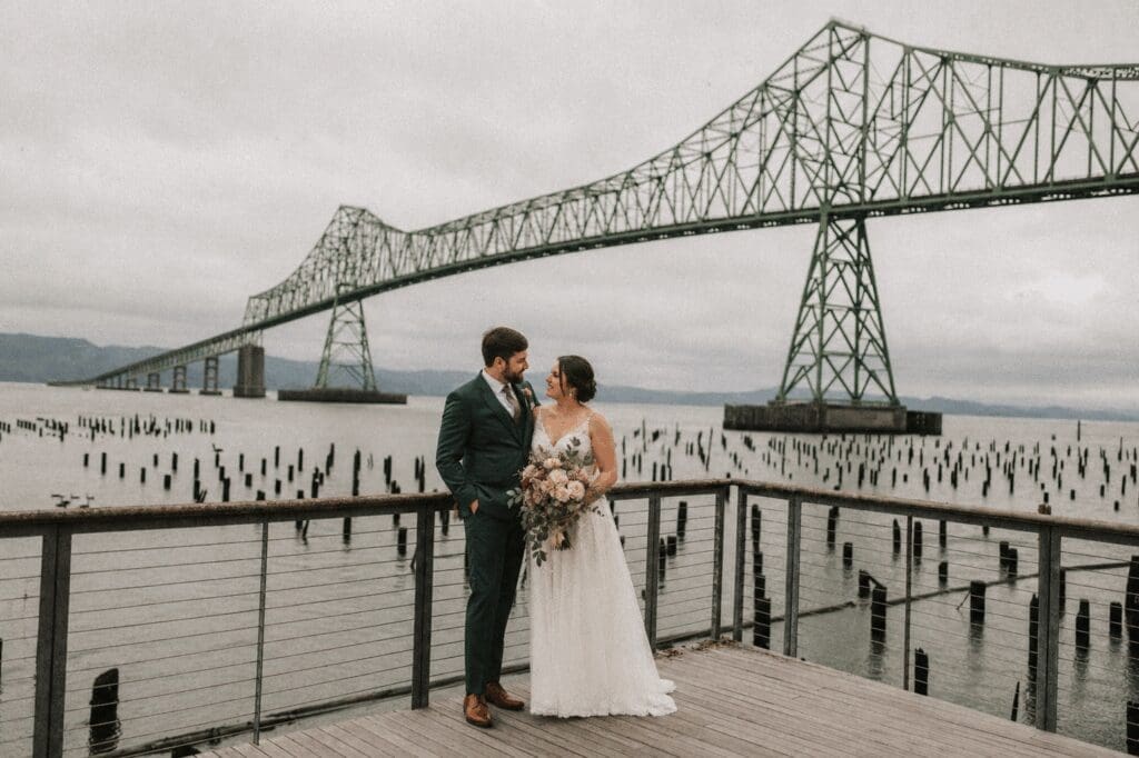 Bride and groom stand on a waterfront deck beneath the Astoria bridge, celebrating their Oregon Coast wedding while a day-of coordinator manages coastal logistics and timeline behind the scenes.