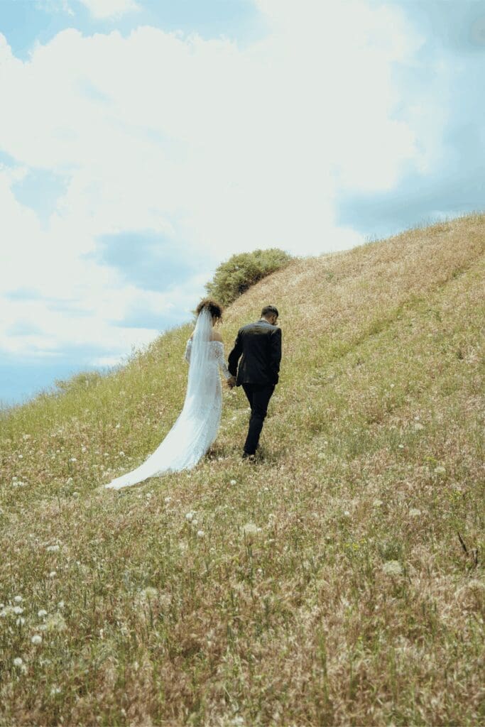 Bride and groom walk hand in hand up a grassy hillside with ocean sky in the distance, symbolizing how an Oregon Coast wedding planner guides couples through permits, travel, and coastal logistics.