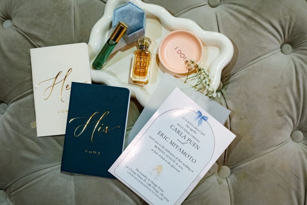 Wedding detail flat lay with his and hers vow books, Carla and Eric's invitation, and perfume before the Lake Oswego ceremony