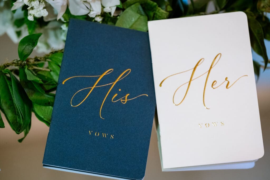 His and Her gold-lettered vow books resting on greenery before the Lake Oswego ceremony