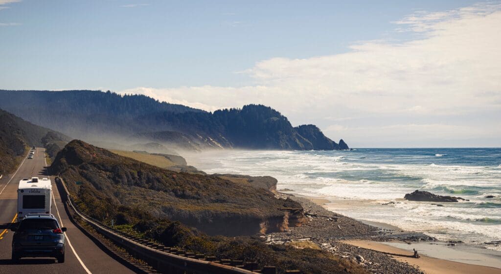 Cars traveling along the Oregon Coast near Lincoln City, with ocean waves, forested cliffs, and a scenic coastal highway.